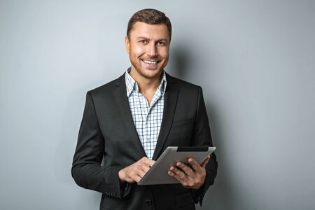 Studio shot of handsome young businessman on grey background. Man wearing jacket and shirt. Man smiling and holding tablet computerの写真素材