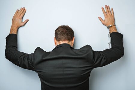 Studio shot of young businessman on grey background. Man wearing jacket. His hands with handcuffs. Back viewの写真素材