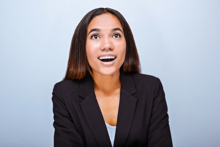 Portrait of beautiful mixed-race young woman standing on grey background. Business woman wearing suit, smiling and wonderingly looking upの写真素材