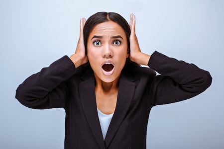 Portrait of beautiful mixed-race young woman standing on grey background. Scared business woman wearing suit and wonderingly looking at camera with hands on earsの写真素材