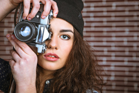Portrait of stylish beautiful young woman with curly hair standing near brick wall. Woman wearing hat and holding vintage cameraの写真素材