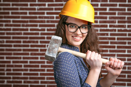 Portrait of stylish beautiful young woman with curly hair standing near brick wall. Woman wearing helmet and glasses. Smiling woman holding big hammerの写真素材