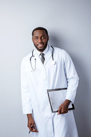 Portrait of male afro american doctor with stethoscope and lab coat. Young doctor with insurance form and looking at camera. Man standing on grey backgroundの写真素材