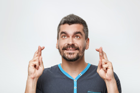Portrait of handsome caucasian man with grey beard standing on white background. Man looking at camera and praying with crossed fingersの写真素材