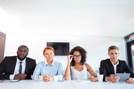 Photo of creative multi ethnic business group. Mixed race business team or commission with electronic devices seriously looking at camera while judging. White modern office interiorの写真素材