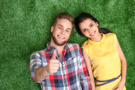 Colorful photo of nice couple lying on green grass. Young man and woman cheerfully smiling and looking at camera. Young man showing thumb upの写真素材