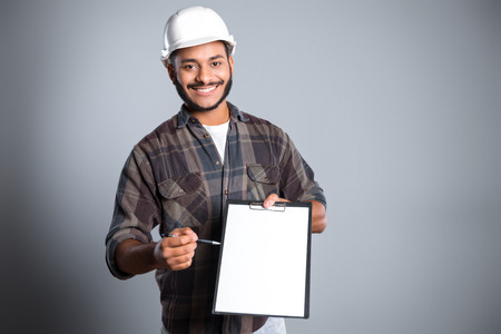 Young mixed race man with beard and helmet standing on grey background. Young builder smiling and proposing to fill formの写真素材