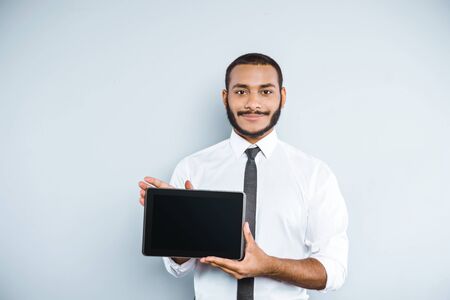Young mixed race businessman with beard standing on grey background. Young man smiling and showing tablet computerの写真素材