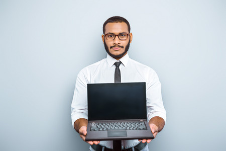 Young mixed race businessman with beard standing on grey background. Young man showing laptopの写真素材