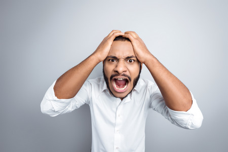 Young mixed race man with beard standing on grey background. Angry young man screaming at cameraの写真素材