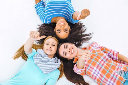 Top view portrait of beautiful three mixed-race girls on white background. Teenagers smiling, showing thumb up and looking at cameraの写真素材