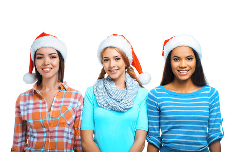 Christmas portrait of beautiful three mixed-race girls. Teenagers wearing santa claus hats. Girls smiling and looking at cameraの写真素材