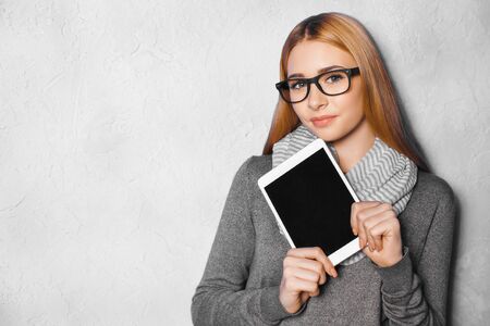 Nice portrait of beautiful girl on grey background. Young woman with glasses holding tablet computer...