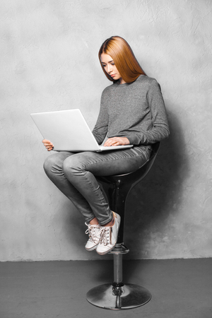 Nice portrait of beautiful girl on grey background. Young woman sitting on high chair and using laptopの写真素材