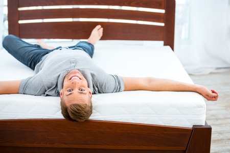 Photo of young man lying on nice white bed. Young man demonstrating quality of mattressの写真素材