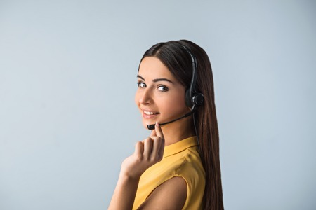 Photo of beautiful young call center operator standing near gray background. Woman with headphones looking at camera and smilingの写真素材