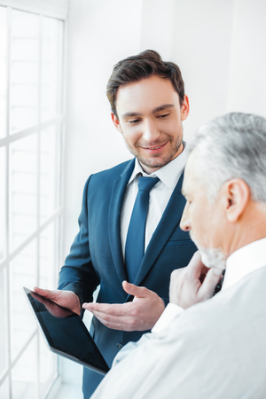 Photo of aged businessman and his colleague. Businessmen working in office with big window. Men using tablet computerの写真素材