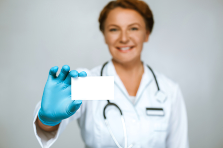 Studio shot of beautiful redheaded female doctor. Medical worker with glove showing visit card. Focus on visit cardの写真素材