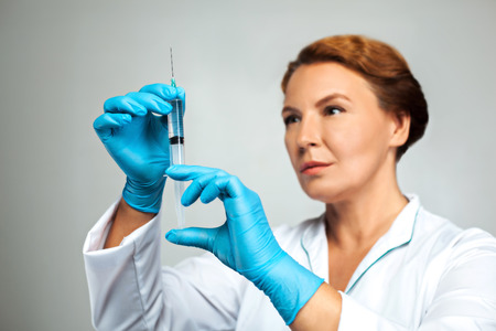 Studio shot of beautiful redheaded female doctor. Medical worker with gloves holding syringe with vaccineの写真素材