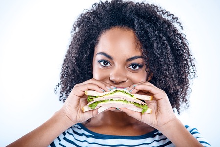 Bright portrait of beautiful young mixed race woman with curly hair on white background. Girl looking at camera and eating sandwichの写真素材