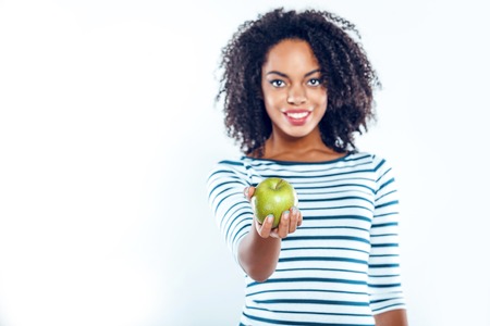 Bright portrait of beautiful young mixed race woman with curly hair on white background. Girl looking at camera, smiling and showing green appleの写真素材