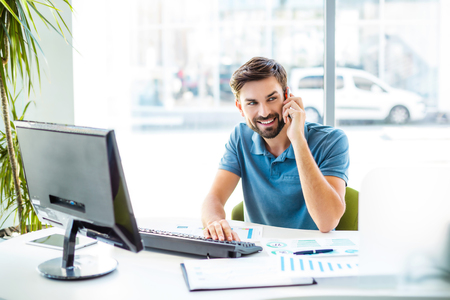Photo of young, handsome, successful and confident businessman. Young businessman working with computer and talking on mobile phone. White modern office interiorの写真素材