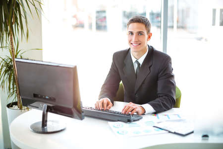 Photo of young and confident businessman. Young businessman working with computer and smiling. White modern office interiorの写真素材