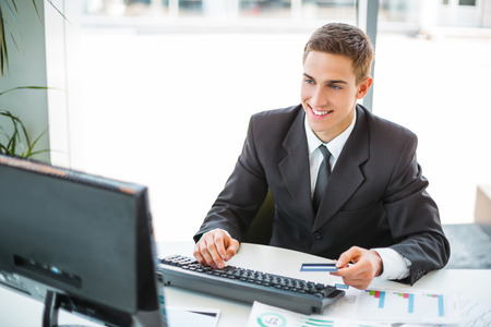 Photo of young and confident businessman. Young businessman working with computer and holding credit card. White modern office interiorの写真素材