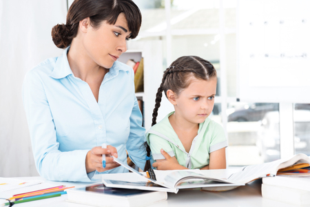 Portrait of cute little girl and her mother while working day. Business woman helping daughter to do homework. Girl is boredの写真素材