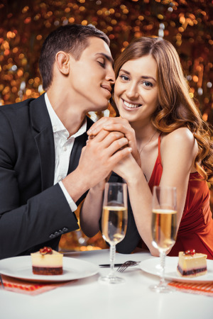 Romantic photo of beautiful couple on glitter gold background. Couple having date at Valentine's Day. Lovers having dinner. There are glasses with champagne and desserts on tableの写真素材