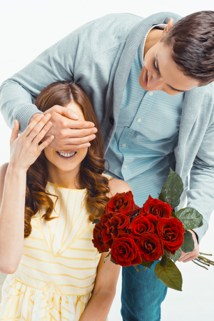 Romantic photo of beautiful couple on white background. Handsome young man giving red roses to his girlfriend and closing her eyesの写真素材
