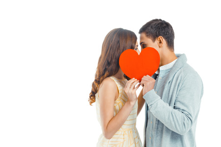 Romantic photo of beautiful couple on white background. Handsome young man and beautiful woman kissing behind Valentine card in shape of heartの写真素材