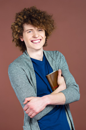 Portrait of stylish handsome young redheaded man. Man standing against brown background, holding book and smilingの写真素材