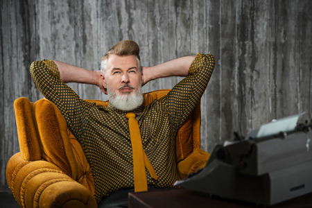 Portrait of stylish handsome writer with beard. Man wearing shirt, sitting at vintage chair near retro typewriter and looking at cameraの写真素材