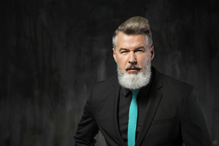 Portrait of stylish handsome adult man with beard. Man wearing black suit with blue tie and looking...