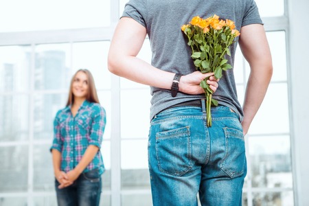 Romantic photo of happy young couple. Young man making surprise for his girlfriend. Man holding bouquet of yellow flowers behind his backの写真素材