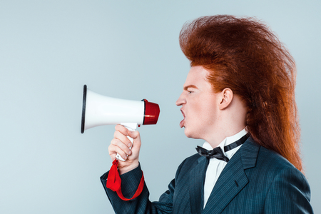 Stylish redheaded young man with bouffant on head. Boy wearing suit with bow-tie and screaming with speakerの写真素材