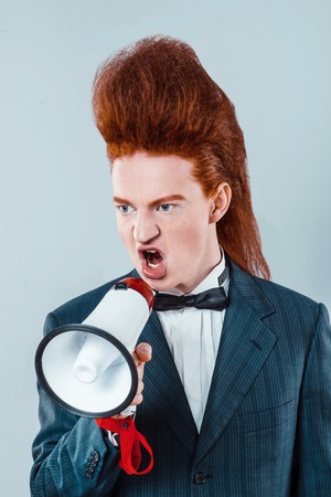 Stylish redheaded young man with bouffant on head. Boy wearing suit with bow-tie and screaming with speakerの写真素材
