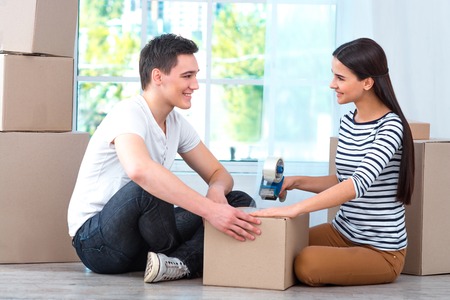 Happy couple smiling, packing and closing carton box, and looking at each otherの写真素材