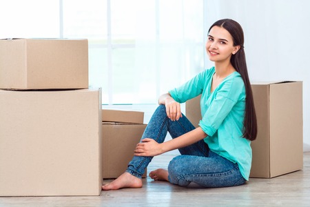Happy woman smiling, packing and closing carton box. Woman sitting on floor near boxes and looking at cameraの写真素材