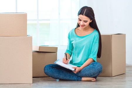 Happy woman smiling, packing and closing carton box. Woman sitting on floor near boxes and holding notebookの写真素材