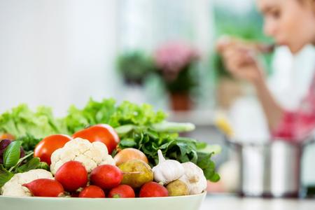 Young blonde woman cooking in kitchen. Woman making healthy vegetable soup. Focus on plate of vegetablesの写真素材