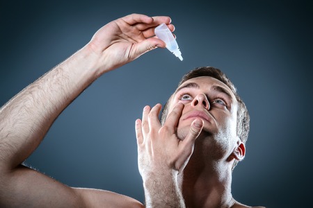 Studio portrait of handsome young man. Clean shaven man with naked torso looking at camera and using eye dropsの写真素材