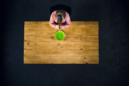 Top view creative photo of girl sitting at wooden vintage table. Girl praying near empty green plateの写真素材