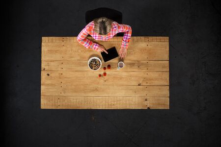 Top view creative photo of girl sitting at wooden vintage table. Girl with plate full of delicious meal. Girl using tablet computerの写真素材