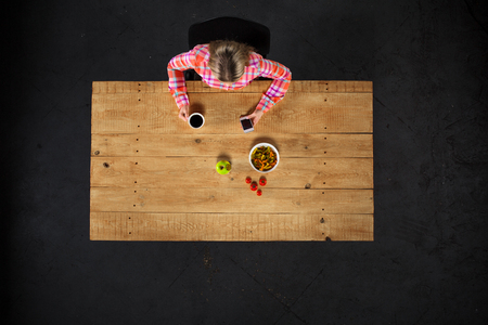 Top view creative photo of girl sitting at wooden vintage table. Girl with plate full of delicious meal. Girl using mobile phoneの写真素材