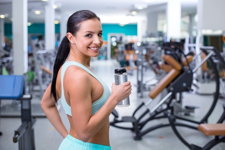 Fitness girl training in sport club with exercise equipments. Woman holding bottle of water, smiling and looking at cameraの写真素材