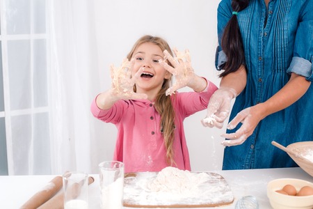 Mother and daughter having fun while preparing meal. Nice white interior. Daughter having hands smeared with doughの写真素材
