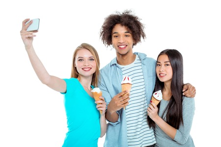 Studio shot of nice young multicultural friends. Beautiful people looking at camera, smiling, making selfie photo on mobile phone and holding ice-creams. Isolated backgroundの写真素材