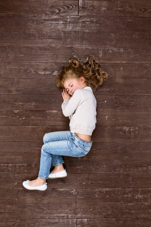 Unhappy child. Top view creative photo of little girl on vintage brown wooden floor. Sad and lonely girl sleepingの写真素材